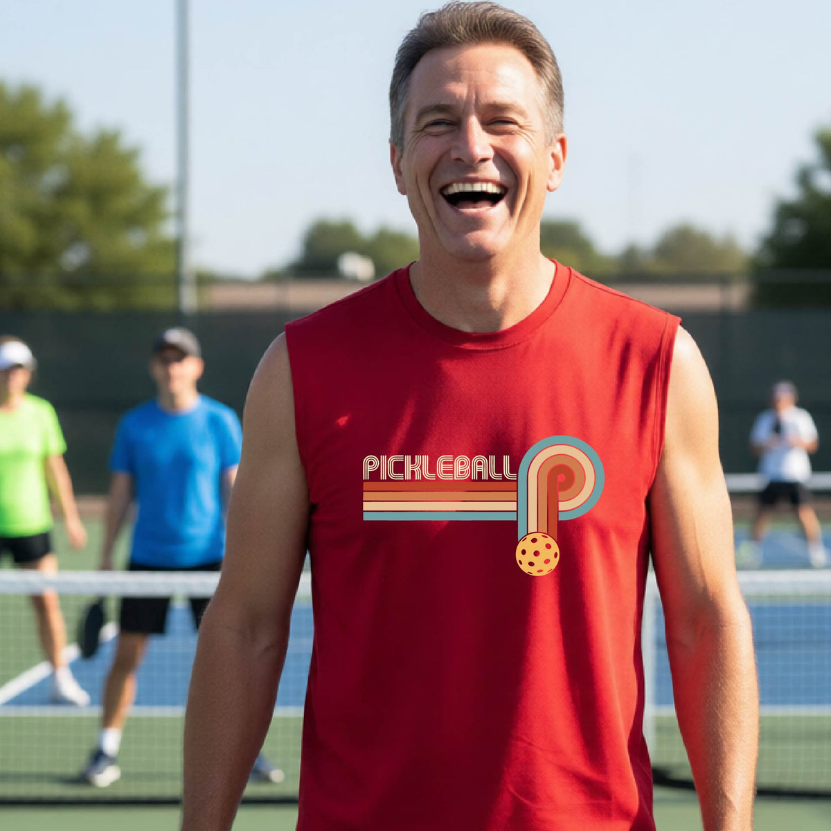 A man on a pickleball court wearing a red 
sleeveless t-shirt with a retro graphic of the word  “pickleball” on top of four colored lines that turn and go down ending in an orange pickleball, standing on a pickleball court.