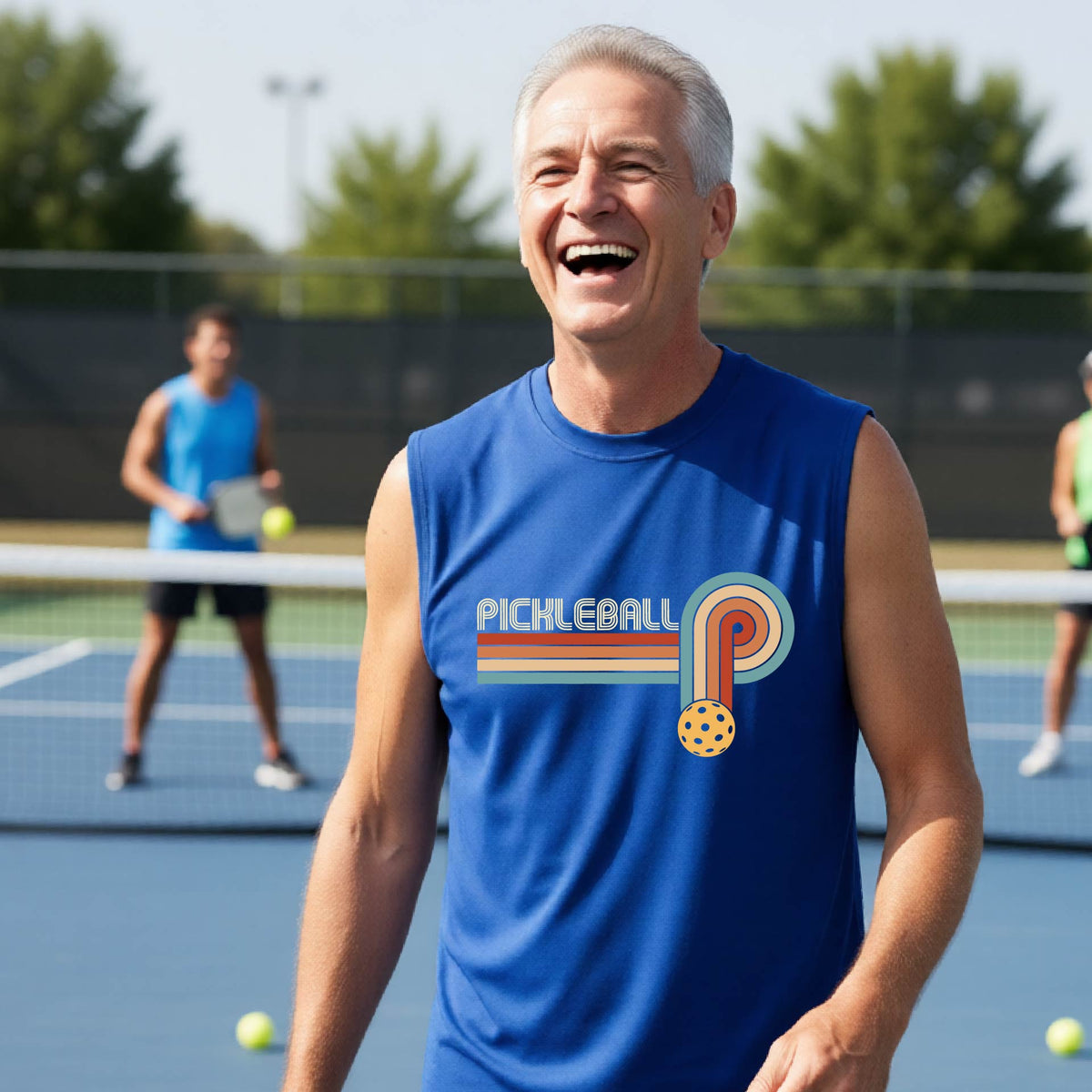 A man on a pickleball court wearing a royal blue sleeveless t-shirt with a retro graphic of the word  “pickleball” on top of four colored lines that turn and go down ending in an orange pickleball, standing on a pickleball court.