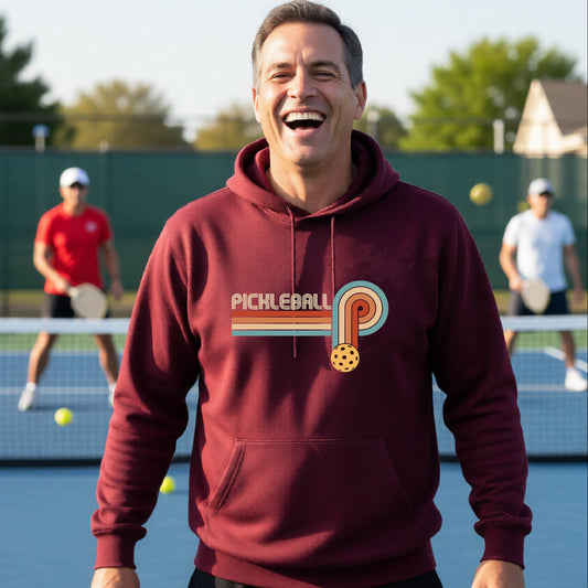 A man on a pickleball court wearing a maroon hoodie pullover with a retro graphic of the word  “pickleball” on top of four colored lines that turn and go down ending in an orange pickleball, standing on a pickleball court.