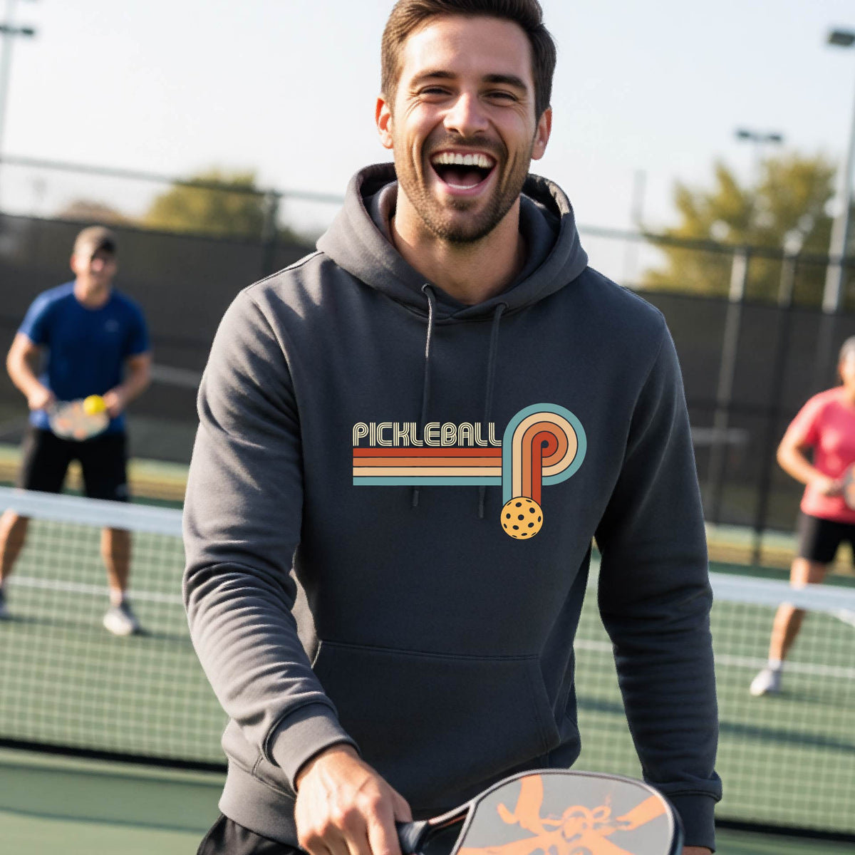 A man on a pickleball court wearing a drk gray hoodie pullover with a retro graphic of the word  “pickleball” on top of four colored lines that turn and go down ending in an orange pickleball, standing on a pickleball court.