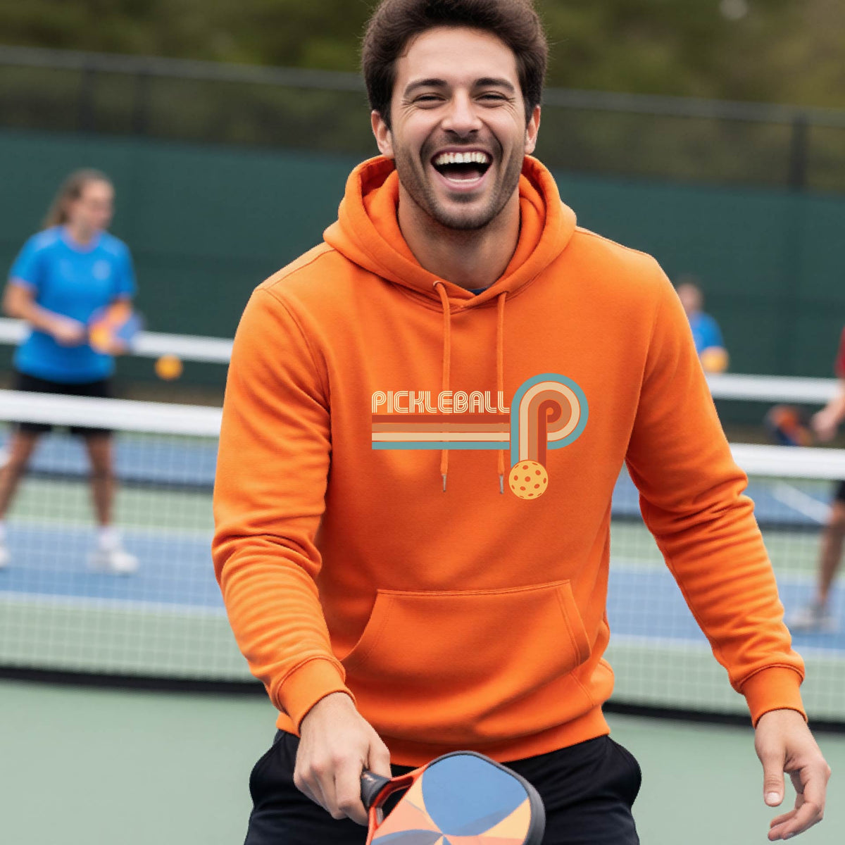 A man on a pickleball court wearing an orange hoodie pullover with a retro graphic of the word  “pickleball” on top of four colored lines that turn and go down ending in an orange pickleball, standing on a pickleball court.