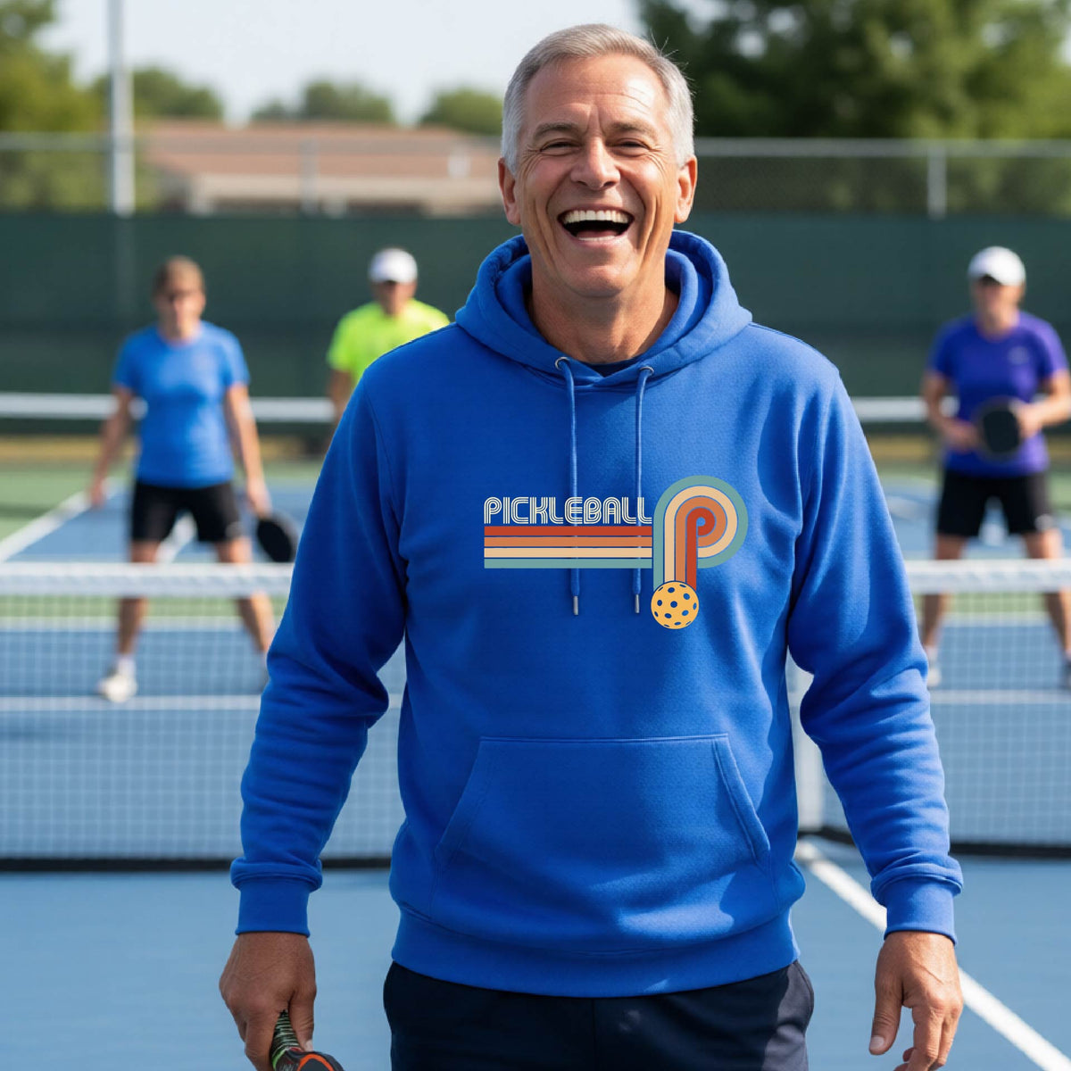 A man on a pickleball court wearing a royal blue hoodie pullover with a retro graphic of the word  “pickleball” on top of four colored lines that turn and go down ending in an orange pickleball, standing on a pickleball court.