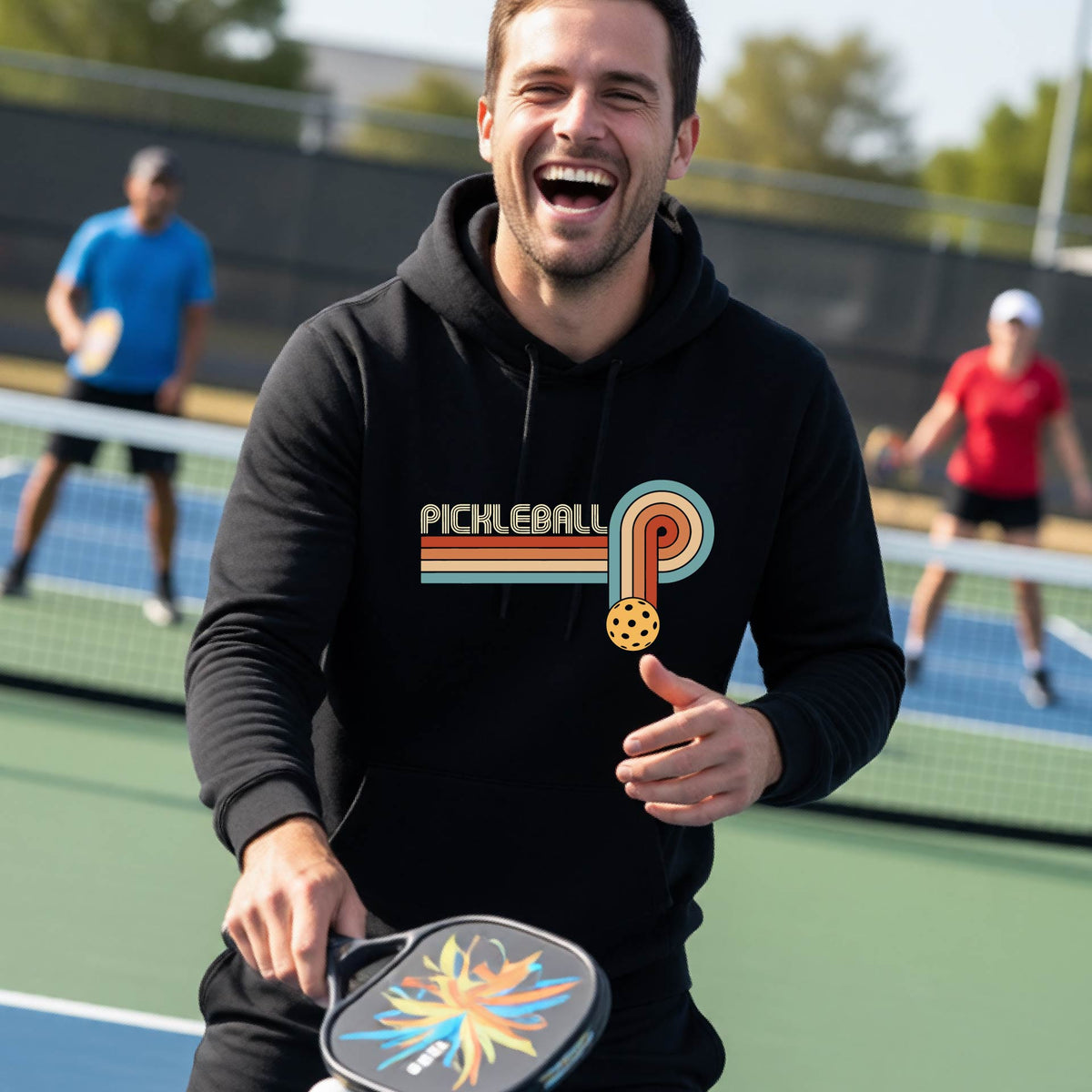 A man on a pickleball court wearing a black hoodie pullover with a retro graphic of the word  “pickleball” on top of four colored lines that turn and go down ending in an orange pickleball, standing on a pickleball court.