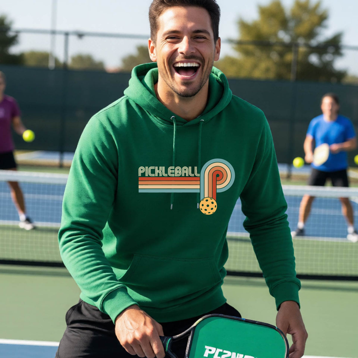 A man on a pickleball court wearing a green hoodie pullover with a retro graphic of the word  “pickleball” on top of four colored lines that turn and go down ending in an orange pickleball, standing on a pickleball court.