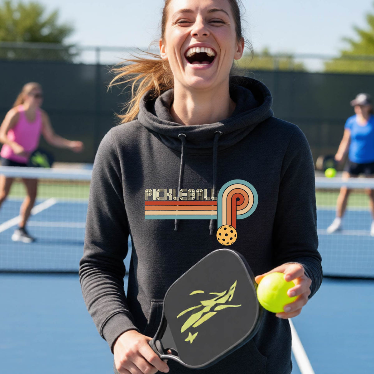 A woman on a pickleball court wearing a black cowl-neck hoodie pullover with a retro graphic of the word  “pickleball” on top of four colored lines that turn and go down ending in an orange pickleball, standing on a pickleball court.