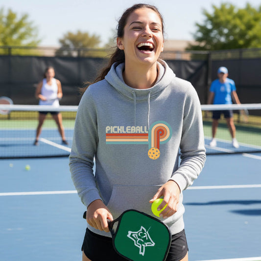 A woman on a pickleball court wearing a gray cowl-neck hoodie pullover with a retro graphic of the word  “pickleball” on top of four colored lines that turn and go down ending in an orange pickleball, standing on a pickleball court.