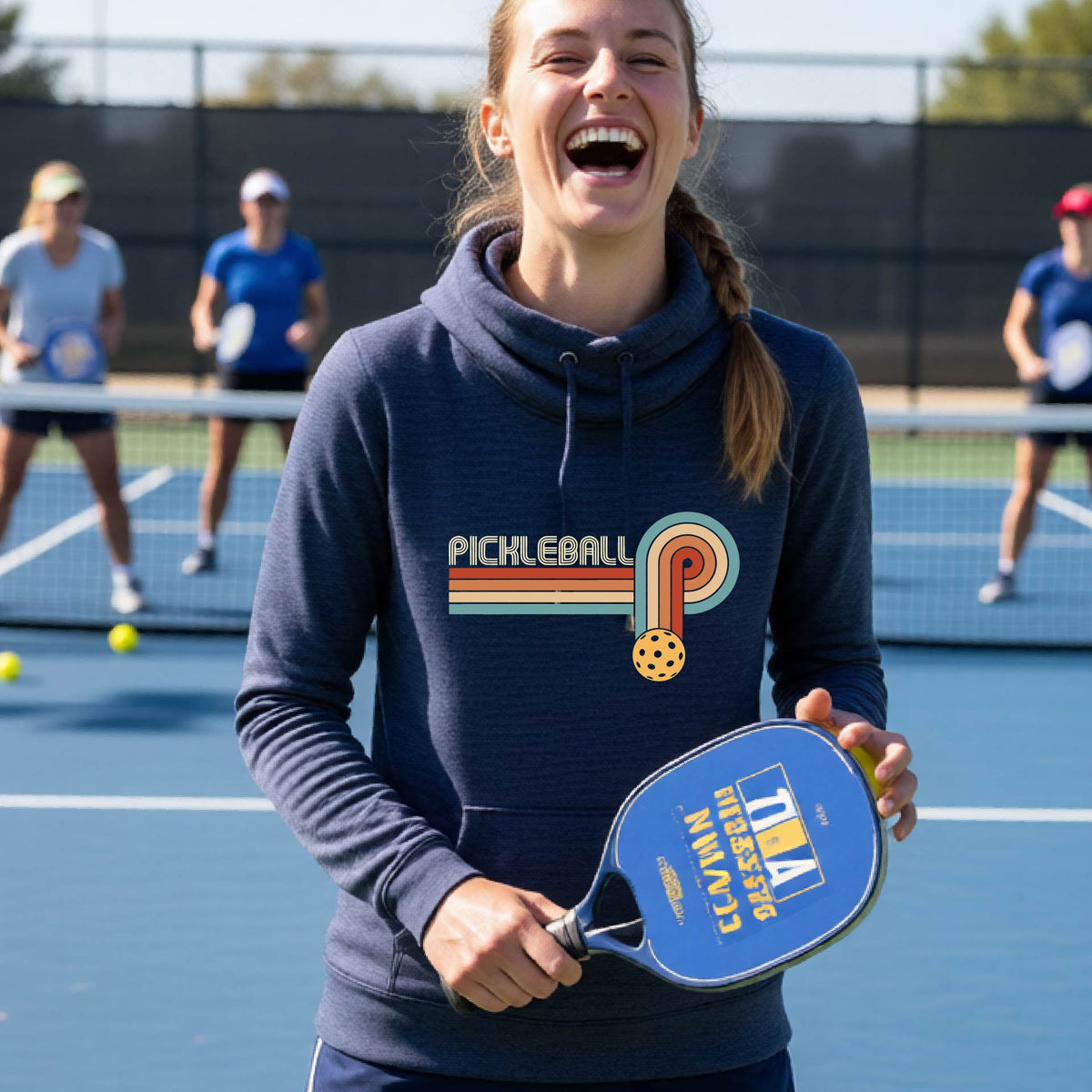 A woman on a pickleball court wearing a navy blue cowl-neck hoodie pullover with a retro graphic of the word  “pickleball” on top of four colored lines that turn and go down ending in an orange pickleball, standing on a pickleball court.