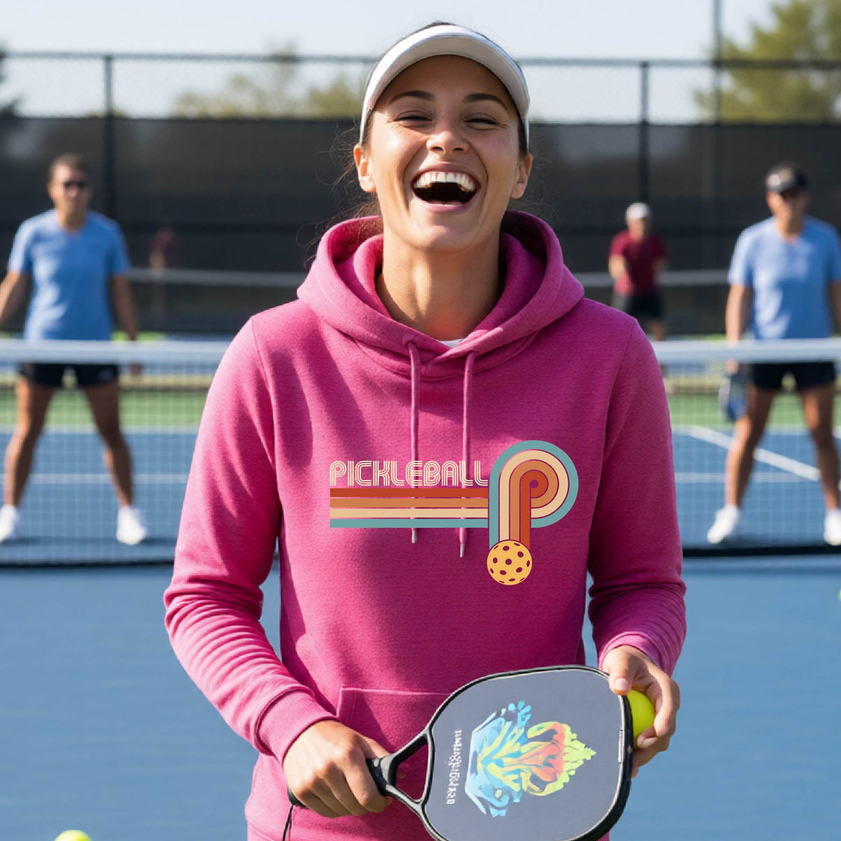 A woman on a pickleball court wearing a pink cowl-neck hoodie pullover with a retro graphic of the word  “pickleball” on top of four colored lines that turn and go down ending in an orange pickleball, standing on a pickleball court.