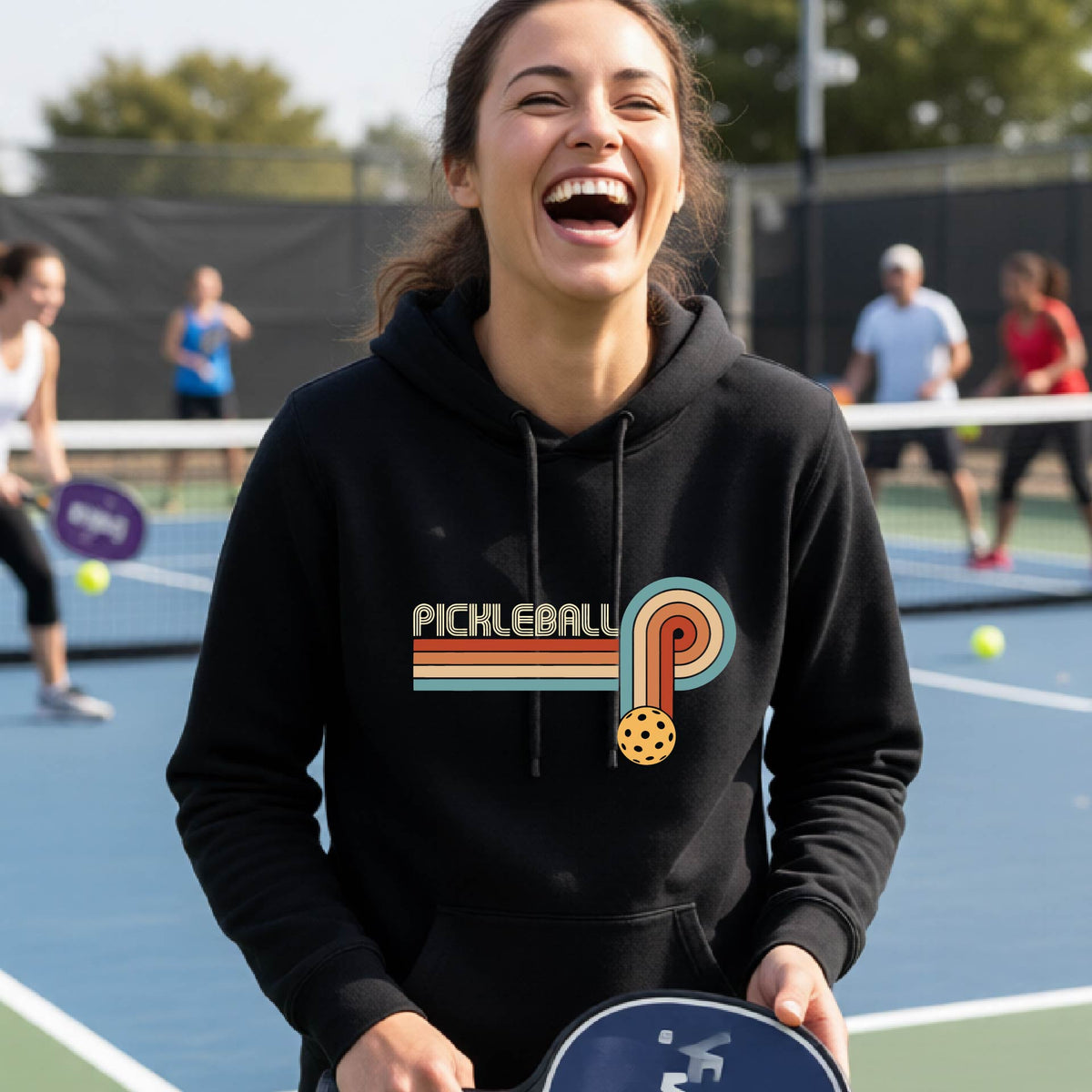 A woman on a pickleball court wearing a black fitted hoodie pullover with a retro graphic of the word  “pickleball” on top of four colored lines that turn and go down ending in an orange pickleball, standing on a pickleball court.