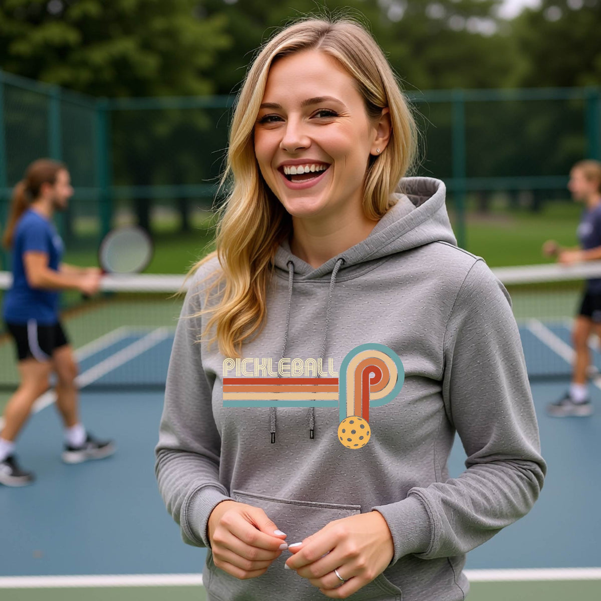 A woman on a pickleball court wearing a gray fitted hoodie pullover with a retro graphic of the word  “pickleball” on top of four colored lines that turn and go down ending in an orange pickleball, standing on a pickleball court.
