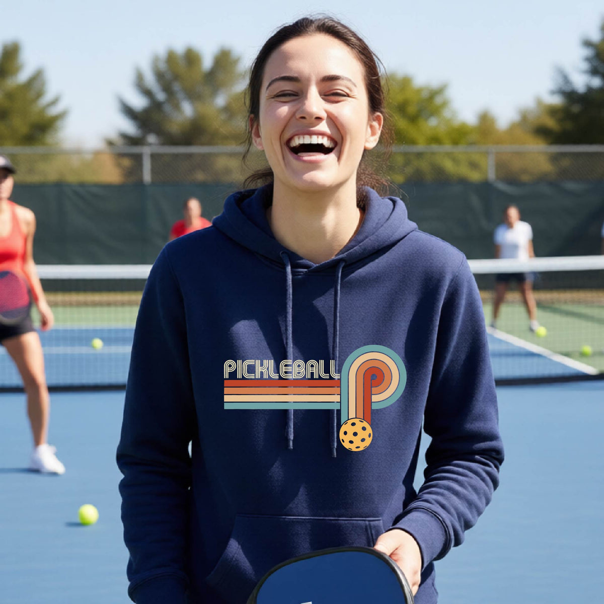 A woman on a pickleball court wearing a navy blue fitted hoodie pullover with a retro graphic of the word  “pickleball” on top of four colored lines that turn and go down ending in an orange pickleball, standing on a pickleball court.