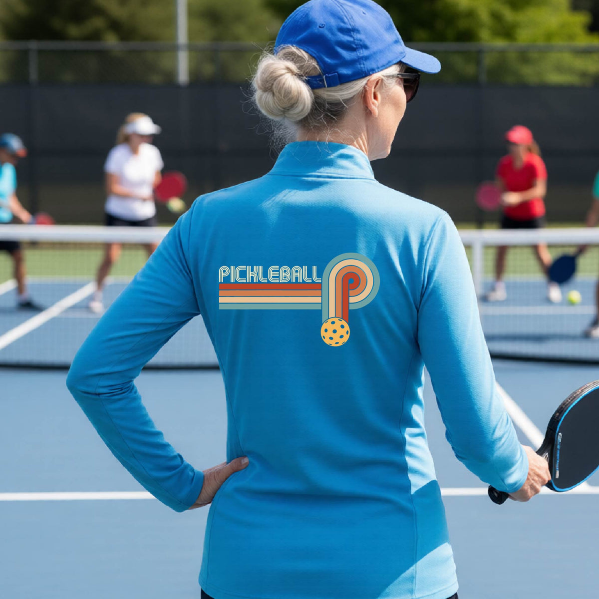 A woman on a pickleball court facing back wearing a bright blue quarter-zip shirt with a retro graphic of the word  “pickleball” on top of four colored lines that turn and go down ending in an orange pickleball, standing on a pickleball court.