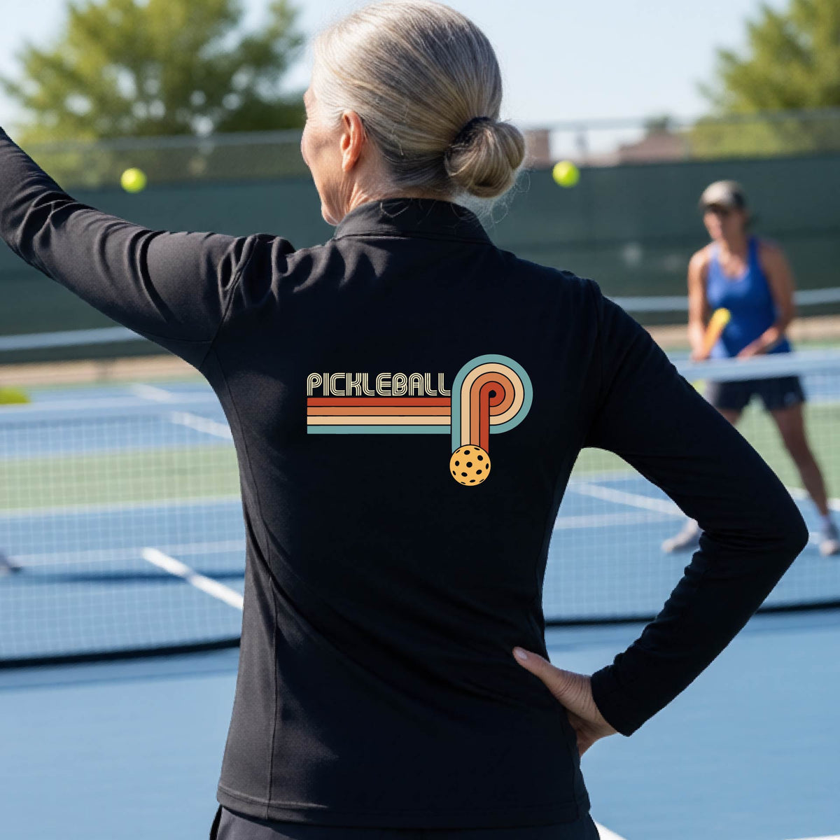 A woman on a pickleball court facing back wearing a black quarter-zip shirt with a retro graphic of the word  “pickleball” on top of four colored lines that turn and go down ending in an orange pickleball, standing on a pickleball court.