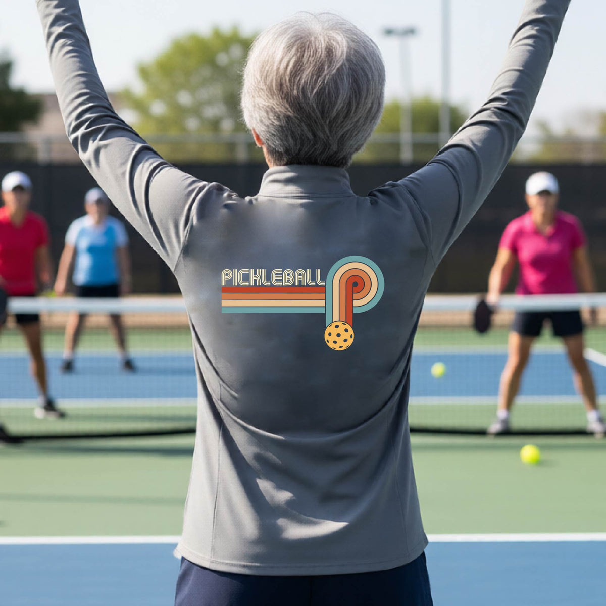 A woman on a pickleball court facing back wearing a gray quarter-zip shirt with a retro graphic of the word  “pickleball” on top of four colored lines that turn and go down ending in an orange pickleball, standing on a pickleball court.