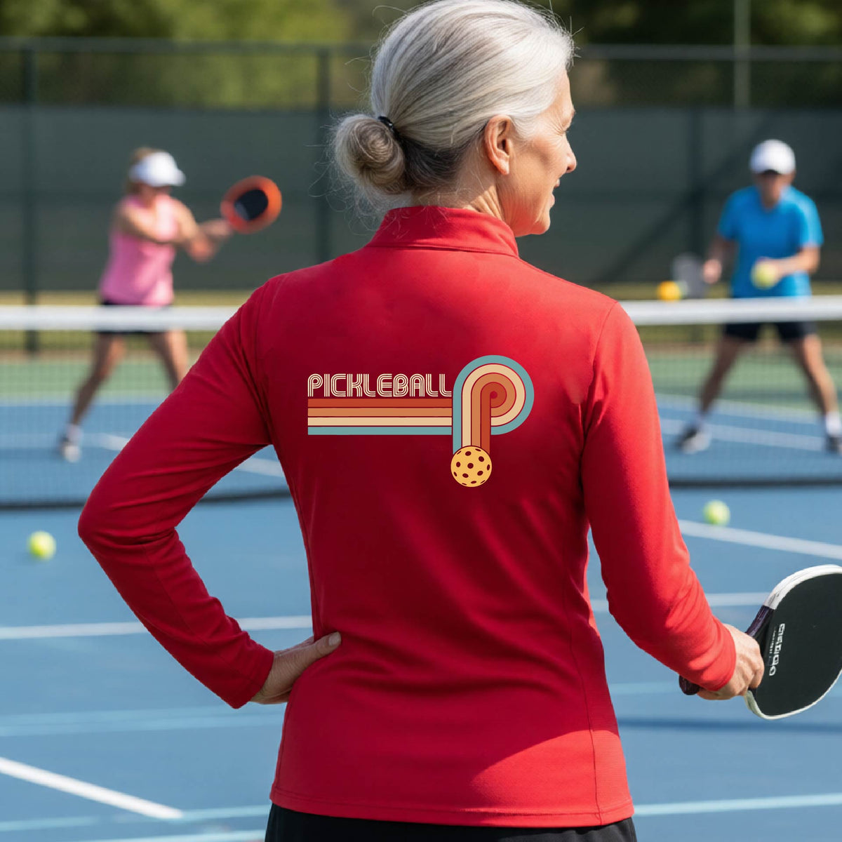 A woman on a pickleball court facing back wearing a red quarter-zip shirt with a retro graphic of the word  “pickleball” on top of four colored lines that turn and go down ending in an orange pickleball, standing on a pickleball court.