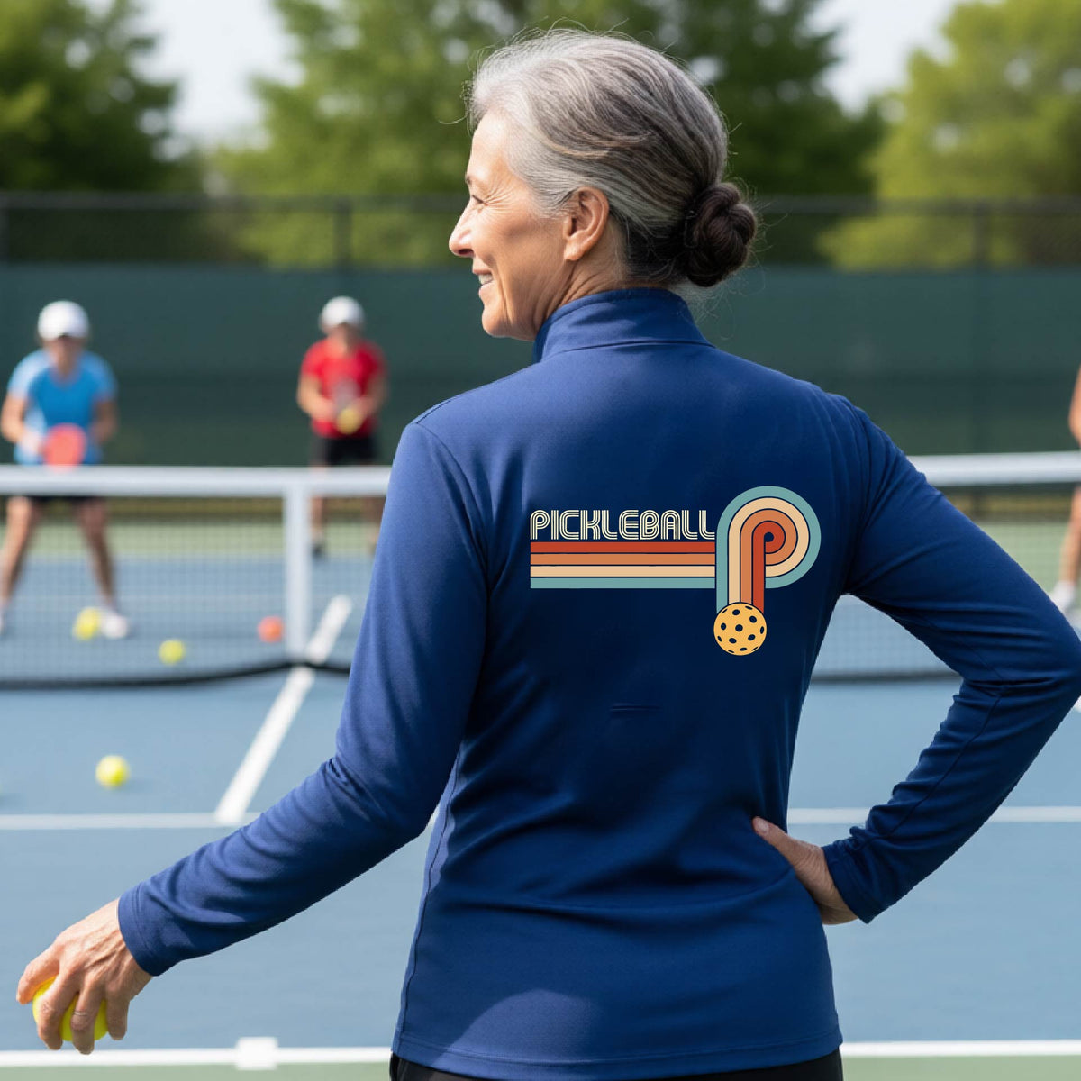 A woman on a pickleball court facing back wearing a royal blue quarter-zip shirt with a retro graphic of the word  “pickleball” on top of four colored lines that turn and go down ending in an orange pickleball, standing on a pickleball court.