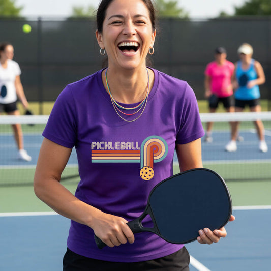 A woman on a pickleball court wearing a purple short-sleeve t-shirt with a retro graphic of the word  “pickleball” on top of four colored lines that turn and go down ending in an orange pickleball, standing on a pickleball court.