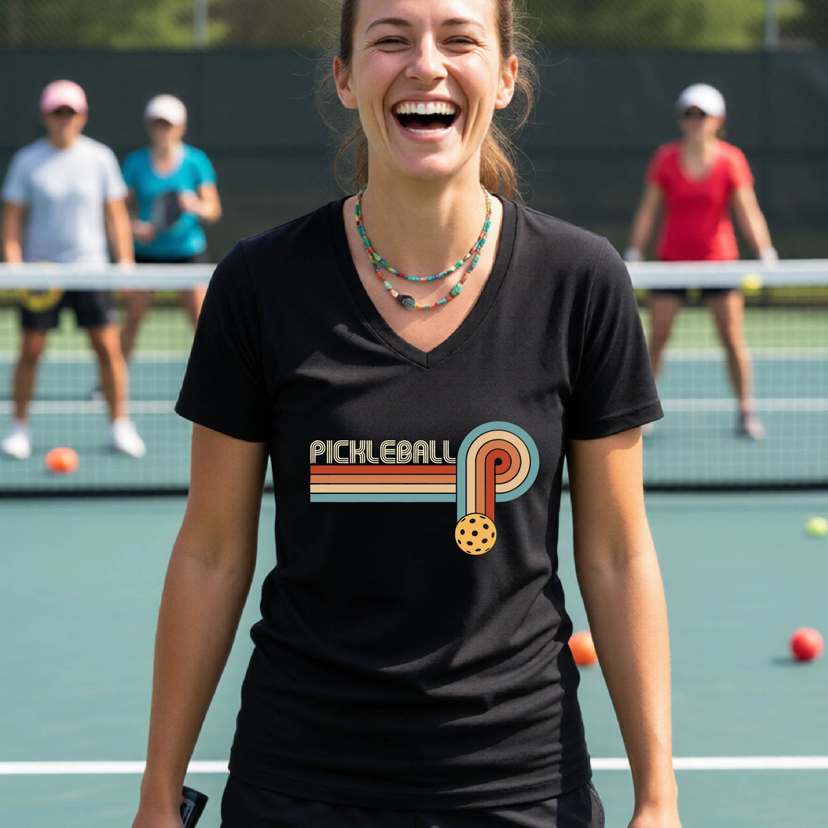 A woman on a pickleball court wearing a black short-sleeve v-neck t-shirt with a retro graphic of the word  “pickleball” on top of four colored lines that turn and go down ending in an orange pickleball, standing on a pickleball court.