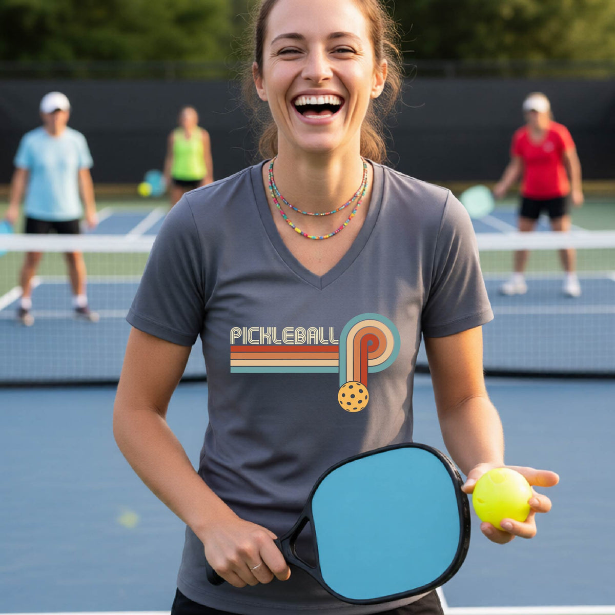 A woman on a pickleball court wearing a gray short-sleeve v-neck t-shirt with a retro graphic of the word  “pickleball” on top of four colored lines that turn and go down ending in an orange pickleball, standing on a pickleball court.