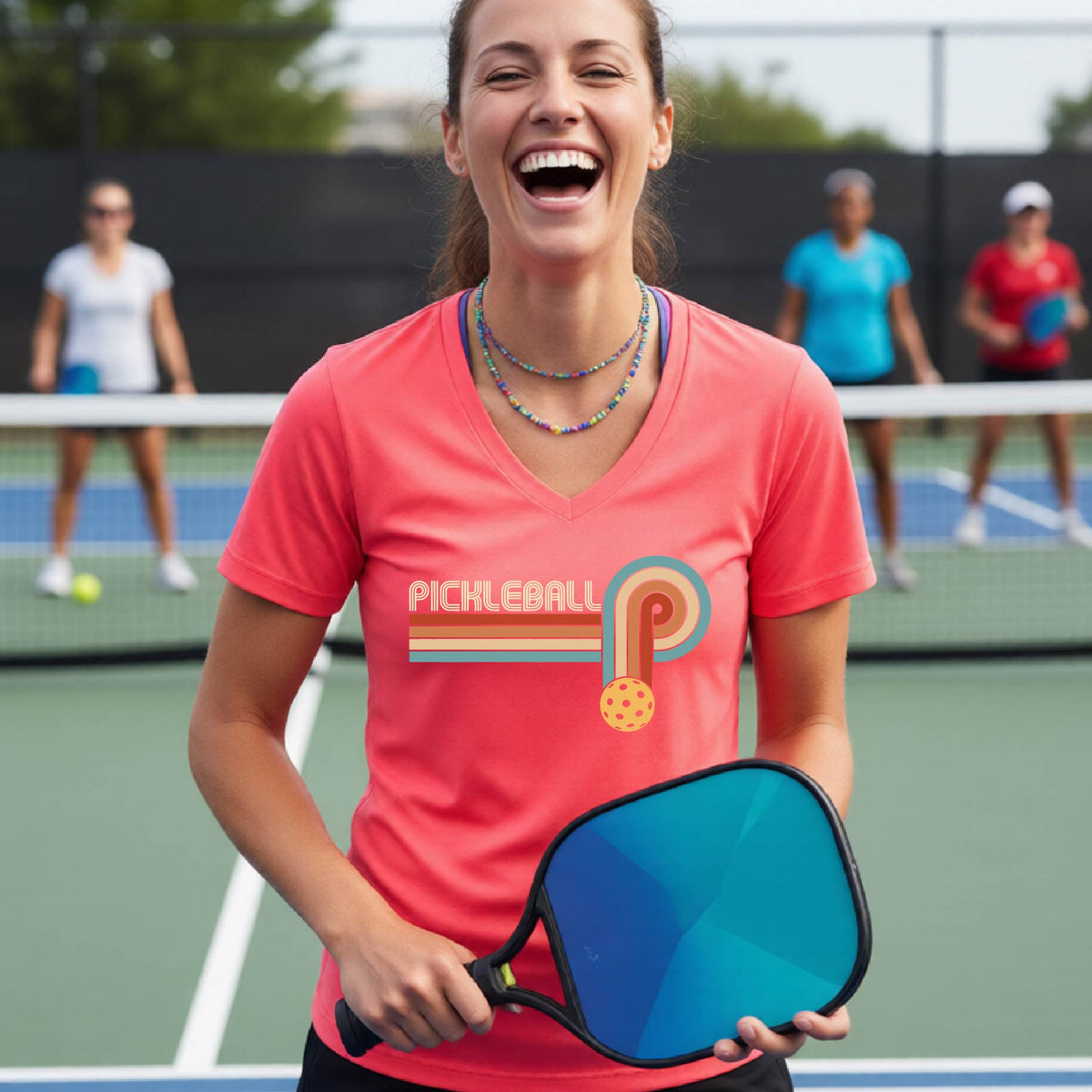 A woman on a pickleball court wearing a coral pink short-sleeve v-neck t-shirt with a retro graphic of the word  “pickleball” on top of four colored lines that turn and go down ending in an orange pickleball, standing on a pickleball court.