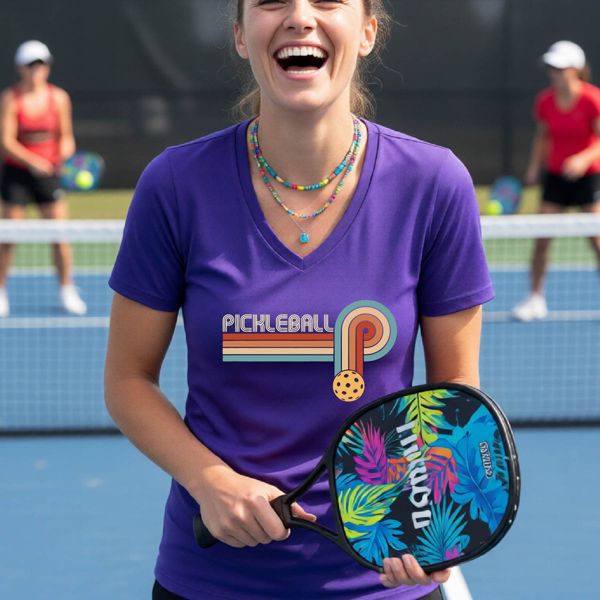 A woman on a pickleball court wearing a purple short-sleeve v-neck t-shirt with a retro graphic of the word  “pickleball” on top of four colored lines that turn and go down ending in an orange pickleball, standing on a pickleball court.