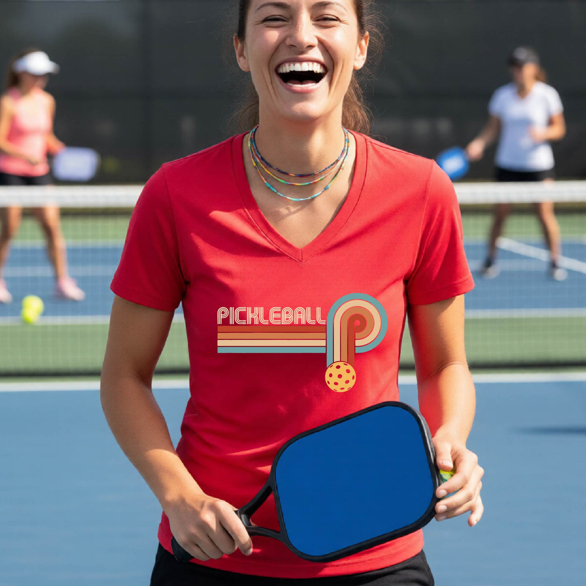 A woman on a pickleball court wearing a red short-sleeve v-neck t-shirt with a retro graphic of the word  “pickleball” on top of four colored lines that turn and go down ending in an orange pickleball, standing on a pickleball court.