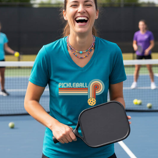 A woman on a pickleball court wearing a black short-sleeve v-neck t-shirt with a retro graphic of the word  “pickleball” on top of four colored lines that turn and go down ending in an orange pickleball, standing on a pickleball court.