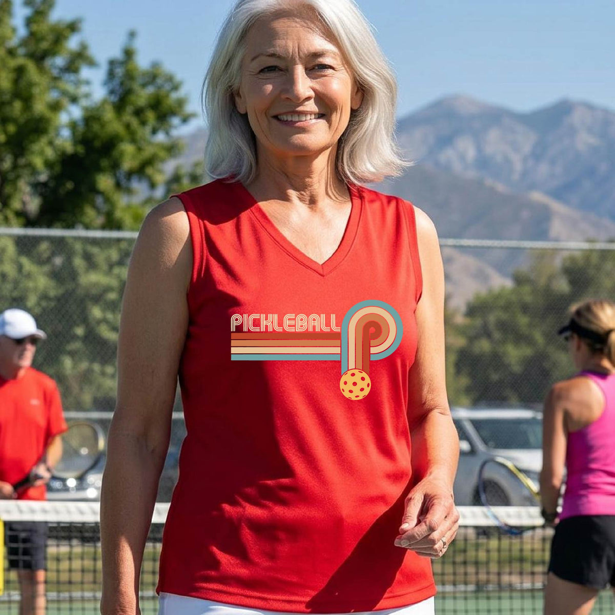 A woman on a pickleball court wearing a red sleeveless t-shirt with a retro graphic of the word  “pickleball” on top of four colored lines that turn and go down ending in an orange pickleball, standing on a pickleball court.