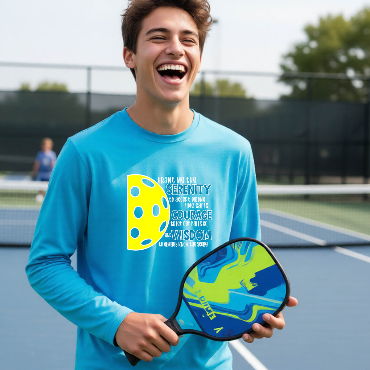 A young person on a pickleball court wearing a bright blue long-sleeve t-shirt with a graphic of the Serenity Prayer made for pickleball:  “Grant me the serenity to Accept Wrong Line Calls; Courage to Let Out Balls Go; and, Wisdom to Always Know the Score” next to a half-pickleball icon, standing on a pickleball court.