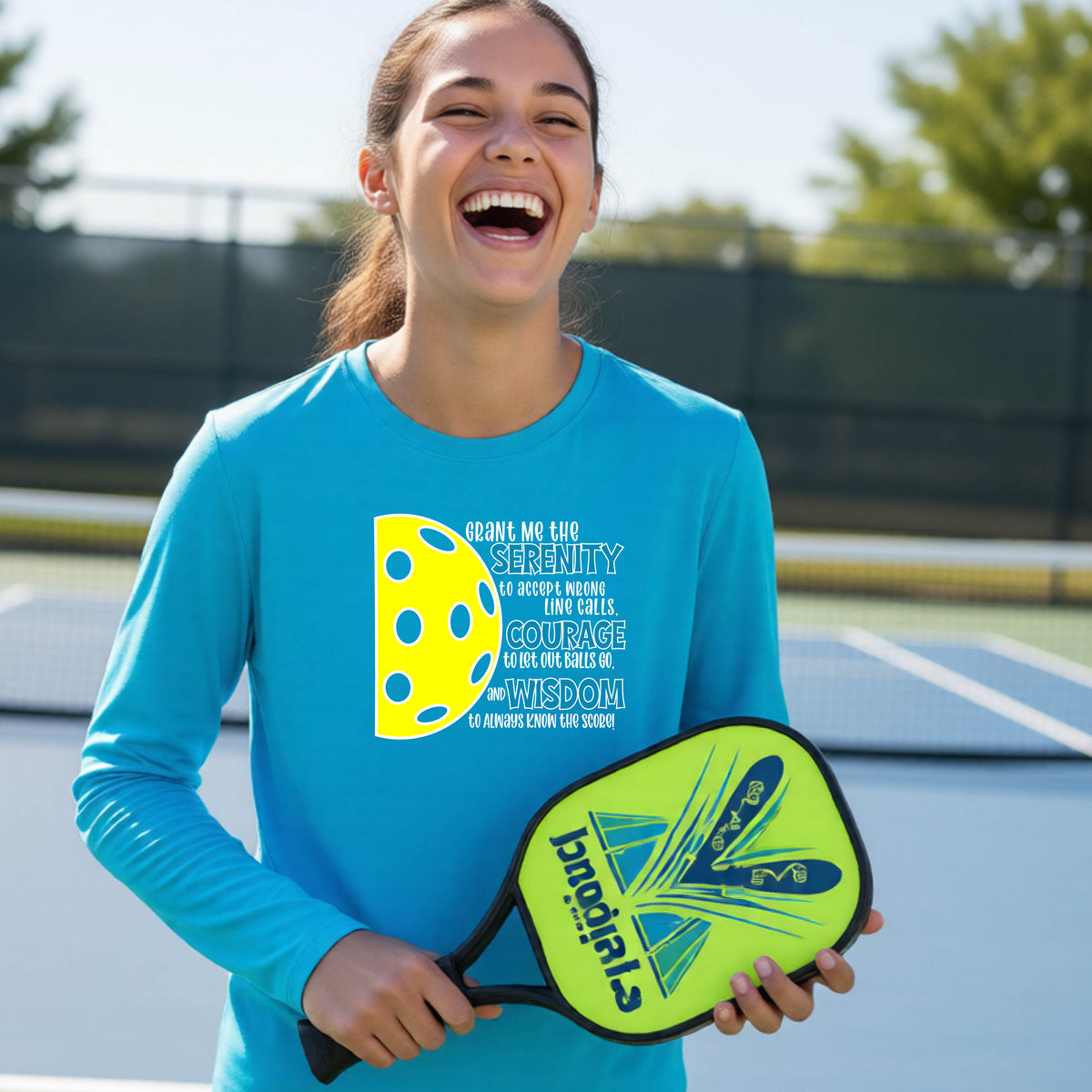 A young person on a pickleball court wearing a bright blue long-sleeve t-shirt with a graphic of the Serenity Prayer made for pickleball:  “Grant me the serenity to Accept Wrong Line Calls; Courage to Let Out Balls Go; and, Wisdom to Always Know the Score” next to a half-pickleball icon, standing on a pickleball court.