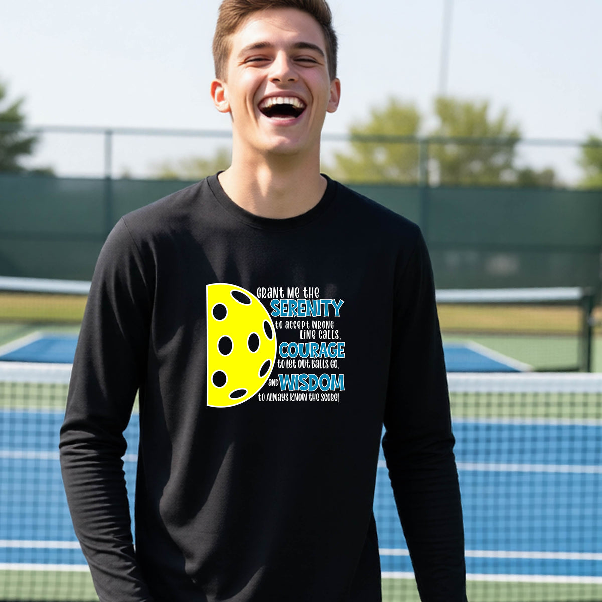A young person on a pickleball court wearing a black long-sleeve t-shirt with a graphic of the Serenity Prayer made for pickleball:  “Grant me the serenity to Accept Wrong Line Calls; Courage to Let Out Balls Go; and, Wisdom to Always Know the Score” next to a half-pickleball icon, standing on a pickleball court.