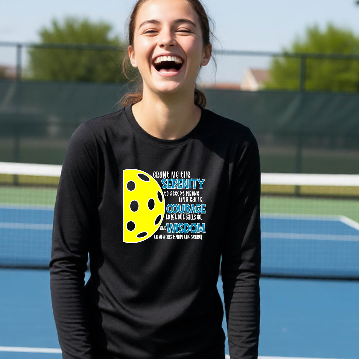 A young person on a pickleball court wearing a black long-sleeve t-shirt with a graphic of the Serenity Prayer made for pickleball:  “Grant me the serenity to Accept Wrong Line Calls; Courage to Let Out Balls Go; and, Wisdom to Always Know the Score” next to a half-pickleball icon, standing on a pickleball court.