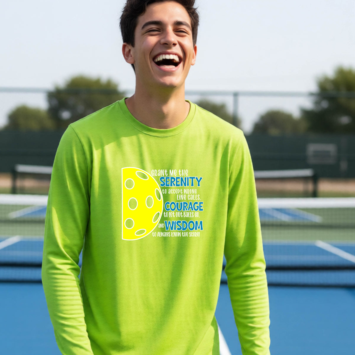 A young person on a pickleball court wearing a lime green long-sleeve t-shirt with a graphic of the Serenity Prayer made for pickleball:  “Grant me the serenity to Accept Wrong Line Calls; Courage to Let Out Balls Go; and, Wisdom to Always Know the Score” next to a half-pickleball icon, standing on a pickleball court.