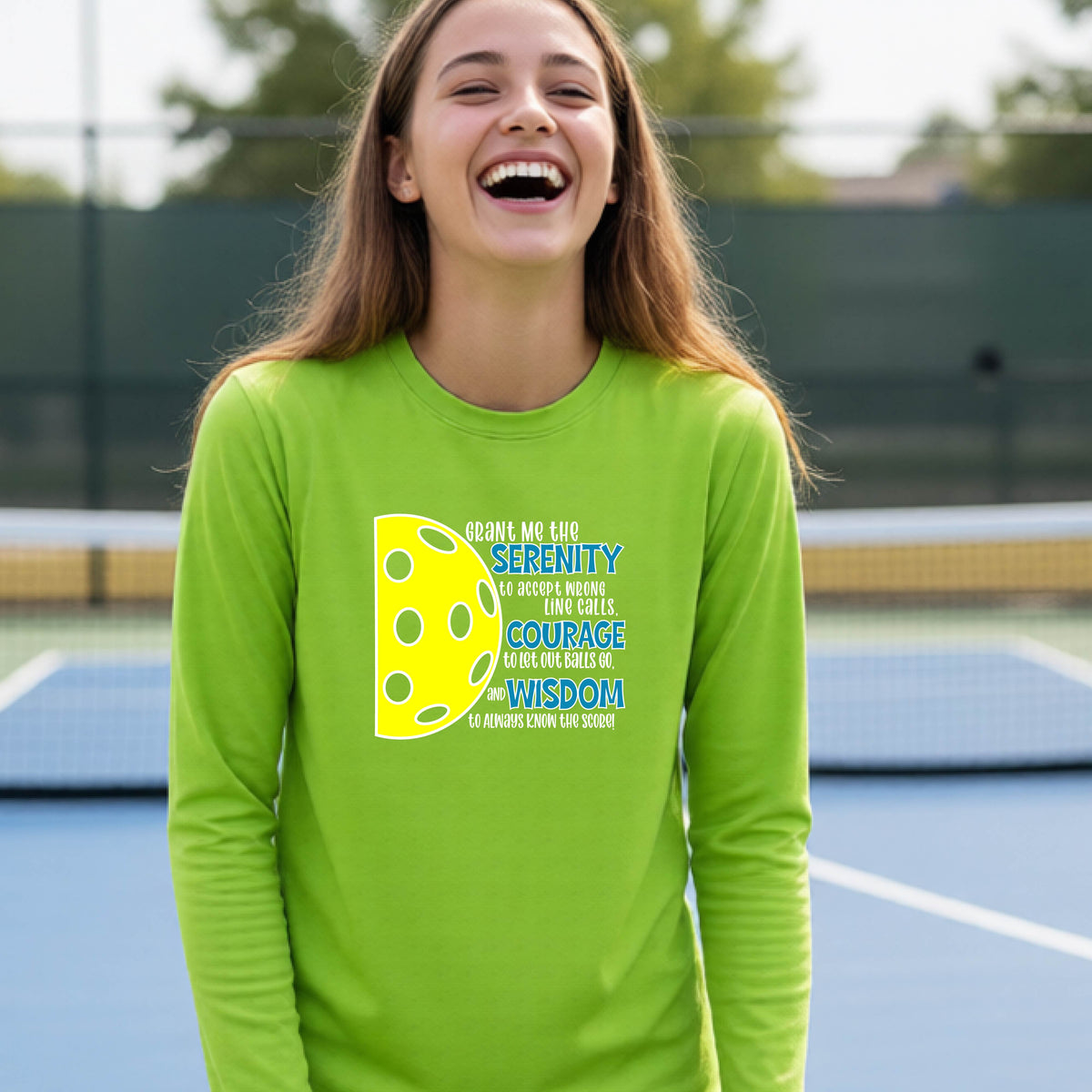 A young person on a pickleball court wearing a lime green long-sleeve t-shirt with a graphic of the Serenity Prayer made for pickleball:  “Grant me the serenity to Accept Wrong Line Calls; Courage to Let Out Balls Go; and, Wisdom to Always Know the Score” next to a half-pickleball icon, standing on a pickleball court.