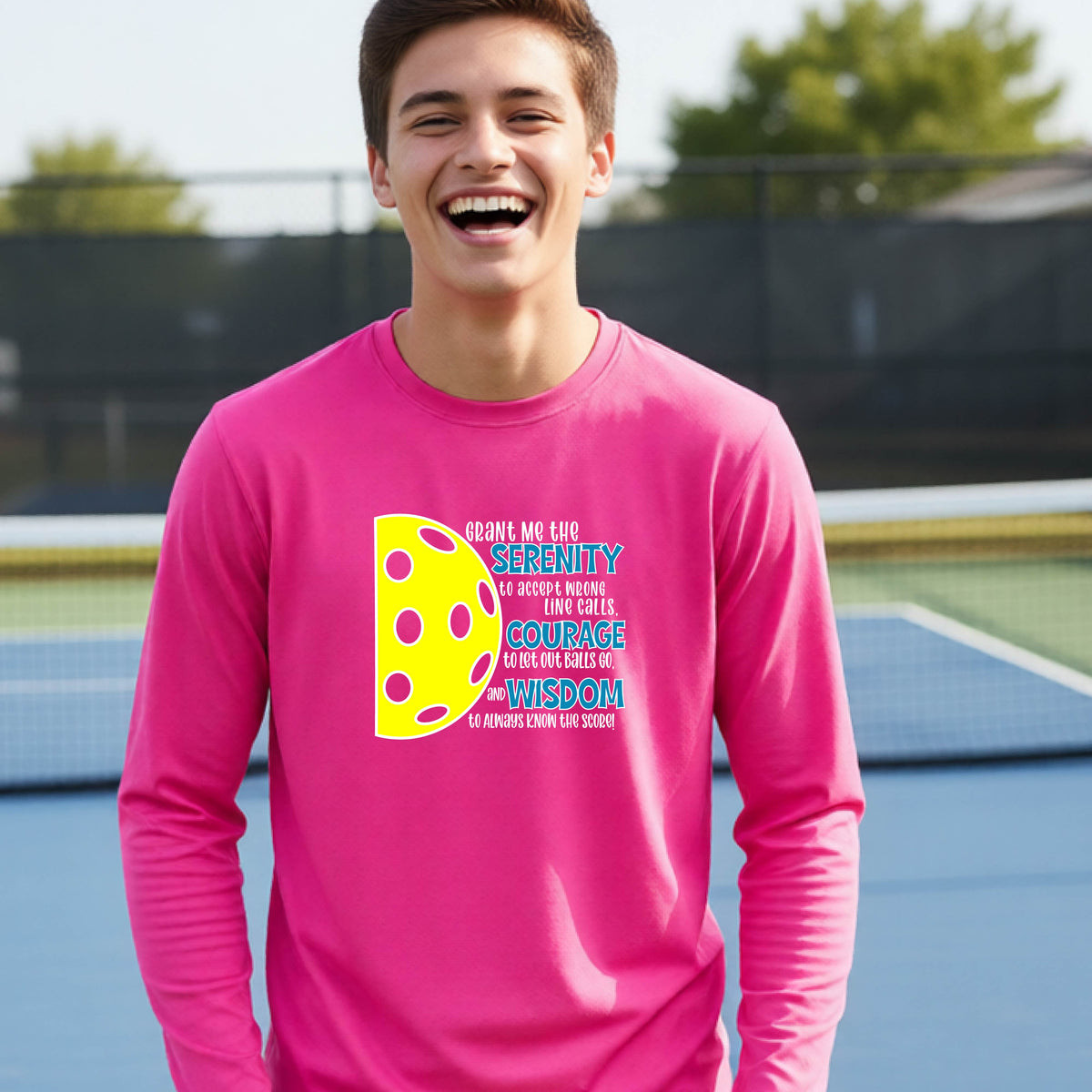 A young person on a pickleball court wearing a pink long-sleeve t-shirt with a graphic of the Serenity Prayer made for pickleball:  “Grant me the serenity to Accept Wrong Line Calls; Courage to Let Out Balls Go; and, Wisdom to Always Know the Score” next to a half-pickleball icon, standing on a pickleball court.