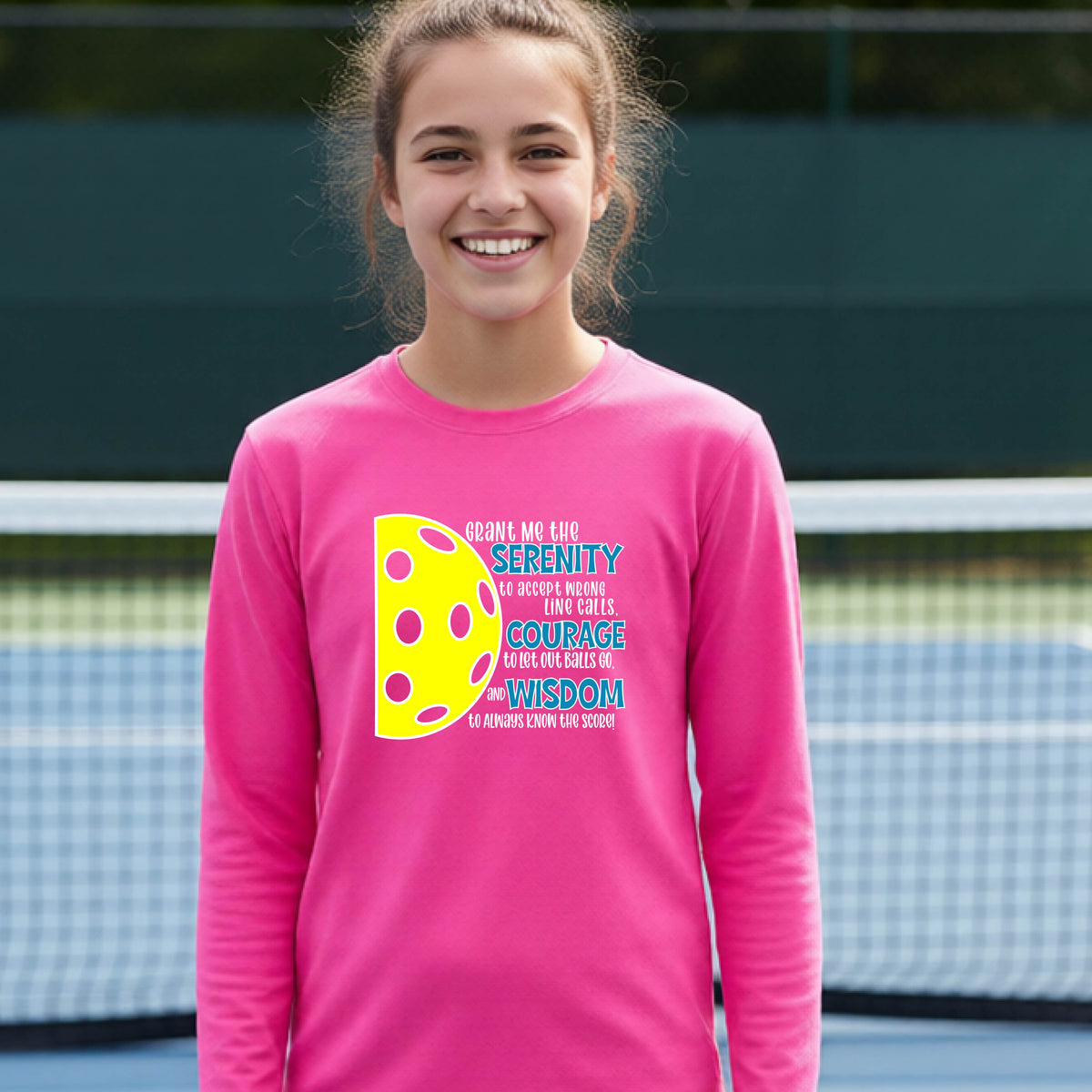 A young person on a pickleball court wearing a pink long-sleeve t-shirt with a graphic of the Serenity Prayer made for pickleball:  “Grant me the serenity to Accept Wrong Line Calls; Courage to Let Out Balls Go; and, Wisdom to Always Know the Score” next to a half-pickleball icon, standing on a pickleball court.