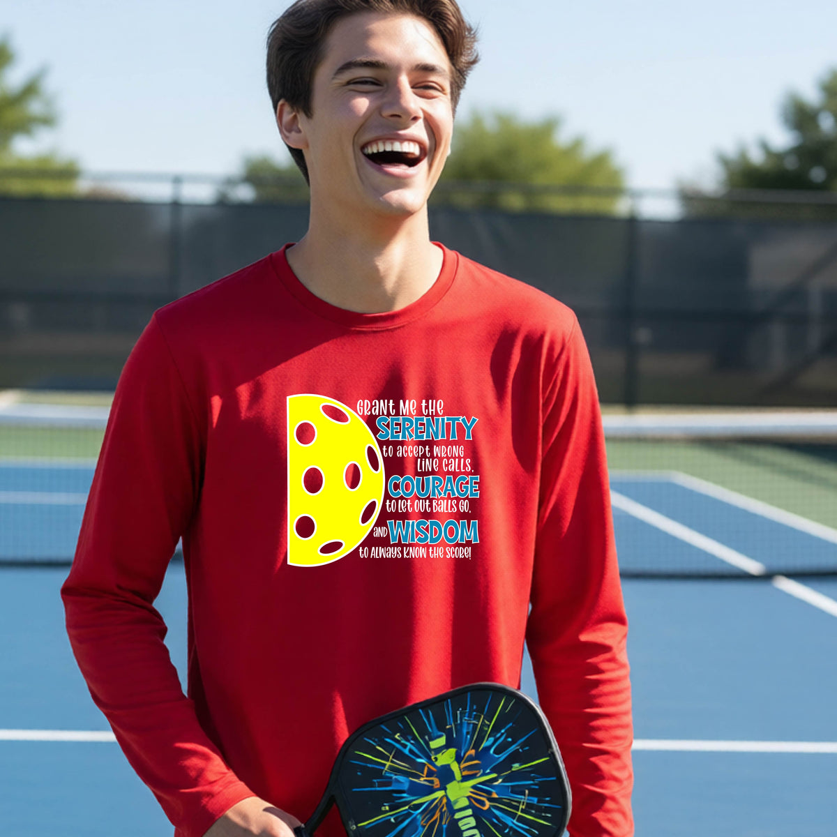 A young person on a pickleball court wearing a red long-sleeve t-shirt with a graphic of the Serenity Prayer made for pickleball:  “Grant me the serenity to Accept Wrong Line Calls; Courage to Let Out Balls Go; and, Wisdom to Always Know the Score” next to a half-pickleball icon, standing on a pickleball court.