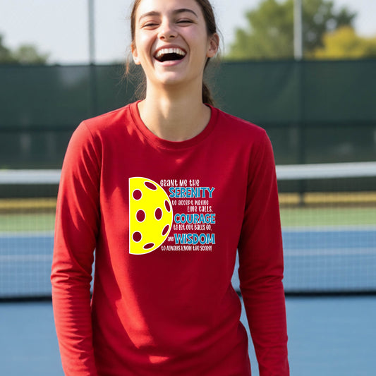 A young person on a pickleball court wearing a  long-sleeve t-shirt with a graphic of the Serenity Prayer made for pickleball:  “Grant me the serenity to Accept Wrong Line Calls; Courage to Let Out Balls Go; and, Wisdom to Always Know the Score” next to a half-pickleball icon, standing on a pickleball court.ed