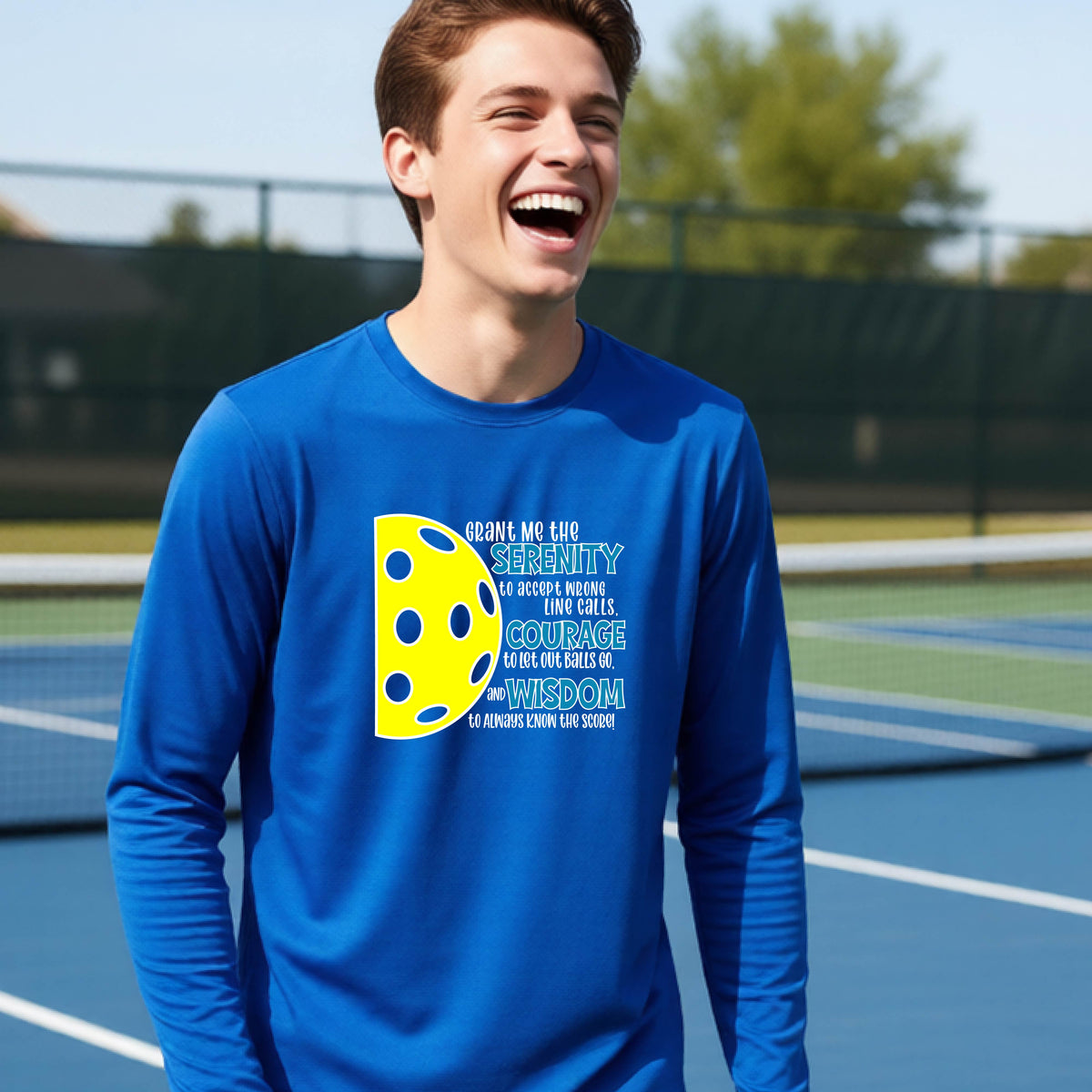 A young person on a pickleball court wearing a royal blue long-sleeve t-shirt with a graphic of the Serenity Prayer made for pickleball:  “Grant me the serenity to Accept Wrong Line Calls; Courage to Let Out Balls Go; and, Wisdom to Always Know the Score” next to a half-pickleball icon, standing on a pickleball court.
