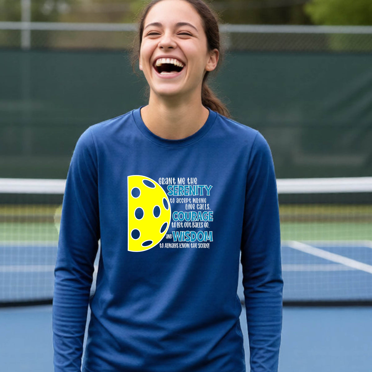 A young person on a pickleball court wearing a royal blue long-sleeve t-shirt with a graphic of the Serenity Prayer made for pickleball:  “Grant me the serenity to Accept Wrong Line Calls; Courage to Let Out Balls Go; and, Wisdom to Always Know the Score” next to a half-pickleball icon, standing on a pickleball court.