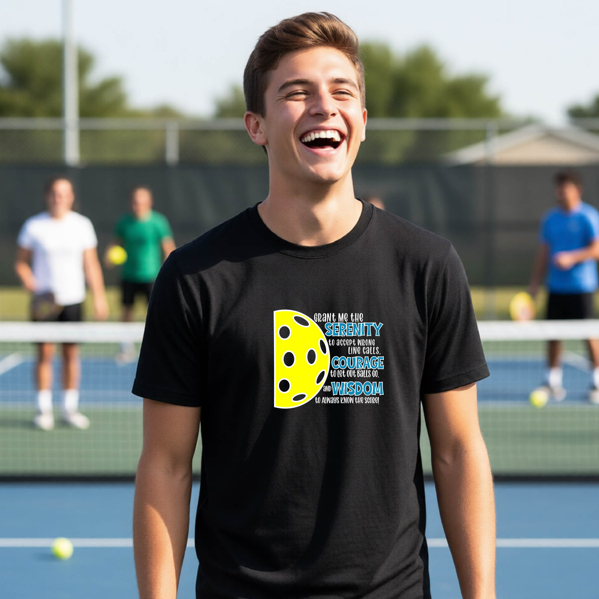 A young person on a pickleball court wearing a black short-sleeve t-shirt with a graphic of the Serenity Prayer made for pickleball:  “Grant me the serenity to Accept Wrong Line Calls; Courage to Let Out Balls Go; and, Wisdom to Always Know the Score” next to a half-pickleball icon, standing on a pickleball court.