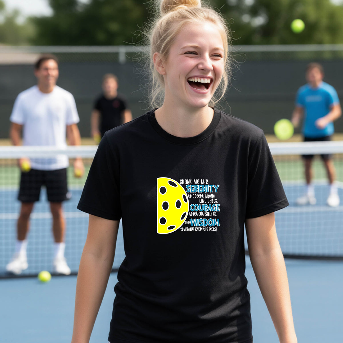 A young person on a pickleball court wearing a black short-sleeve t-shirt with a graphic of the Serenity Prayer made for pickleball:  “Grant me the serenity to Accept Wrong Line Calls; Courage to Let Out Balls Go; and, Wisdom to Always Know the Score” next to a half-pickleball icon, standing on a pickleball court.