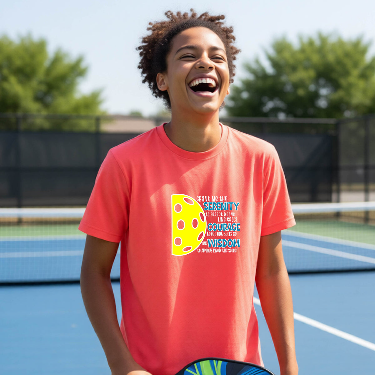 A young person on a pickleball court wearing a coral pink short-sleeve t-shirt with a graphic of the Serenity Prayer made for pickleball:  “Grant me the serenity to Accept Wrong Line Calls; Courage to Let Out Balls Go; and, Wisdom to Always Know the Score” next to a half-pickleball icon, standing on a pickleball court.