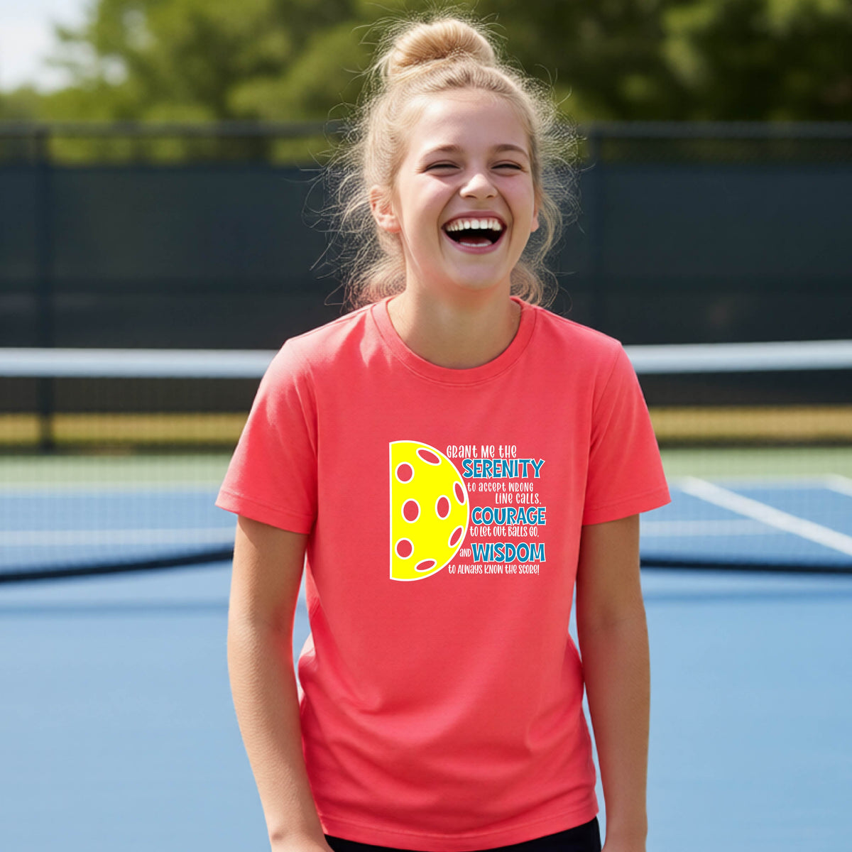 A young person on a pickleball court wearing a coral pink short-sleeve t-shirt with a graphic of the Serenity Prayer made for pickleball:  “Grant me the serenity to Accept Wrong Line Calls; Courage to Let Out Balls Go; and, Wisdom to Always Know the Score” next to a half-pickleball icon, standing on a pickleball court.