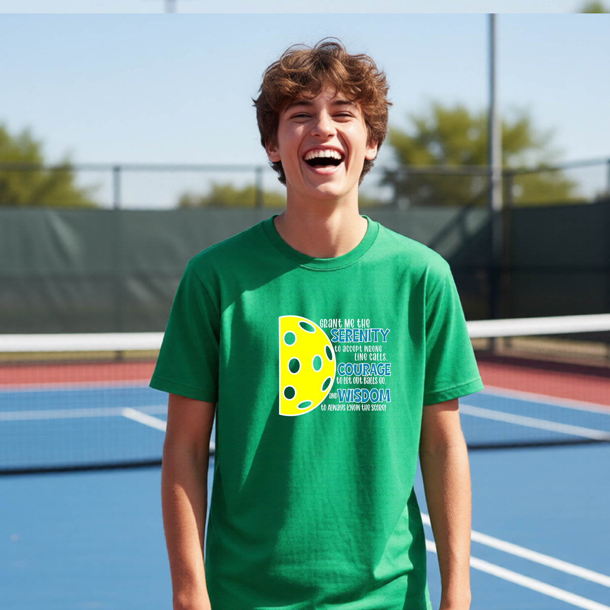 A young person on a pickleball court wearing a green short-sleeve t-shirt with a graphic of the Serenity Prayer made for pickleball:  “Grant me the serenity to Accept Wrong Line Calls; Courage to Let Out Balls Go; and, Wisdom to Always Know the Score” next to a half-pickleball icon, standing on a pickleball court.