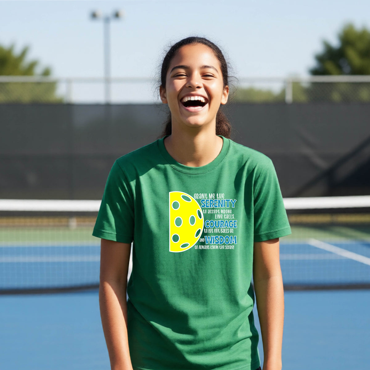 A young person on a pickleball court wearing a green short-sleeve t-shirt with a graphic of the Serenity Prayer made for pickleball:  “Grant me the serenity to Accept Wrong Line Calls; Courage to Let Out Balls Go; and, Wisdom to Always Know the Score” next to a half-pickleball icon, standing on a pickleball court.