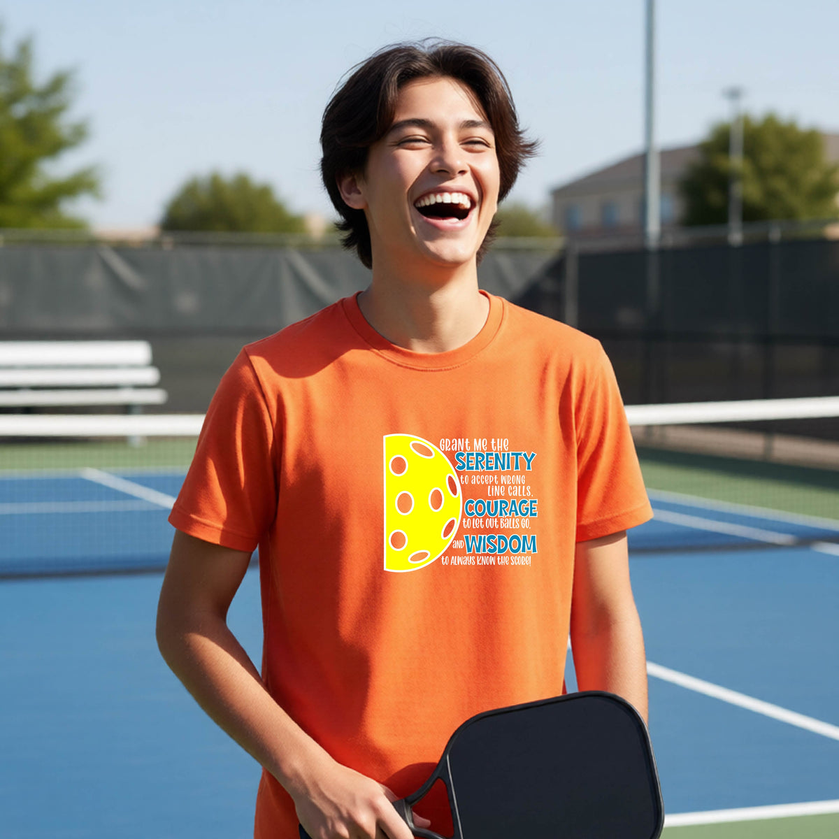 A young person on a pickleball court wearing an orange short-sleeve t-shirt with a graphic of the Serenity Prayer made for pickleball:  “Grant me the serenity to Accept Wrong Line Calls; Courage to Let Out Balls Go; and, Wisdom to Always Know the Score” next to a half-pickleball icon, standing on a pickleball court.