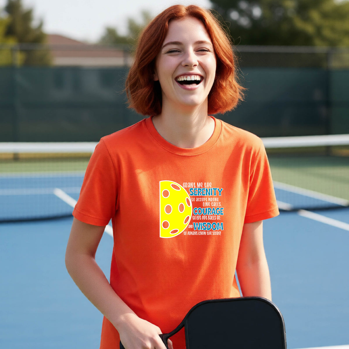 A young person on a pickleball court wearing an orange short-sleeve t-shirt with a graphic of the Serenity Prayer made for pickleball:  “Grant me the serenity to Accept Wrong Line Calls; Courage to Let Out Balls Go; and, Wisdom to Always Know the Score” next to a half-pickleball icon, standing on a pickleball court.