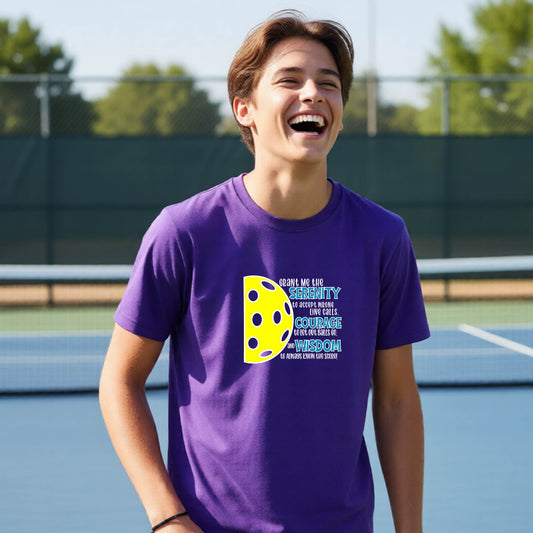 A young person on a pickleball court wearing a purple short-sleeve t-shirt with a graphic of the Serenity Prayer made for pickleball:  “Grant me the serenity to Accept Wrong Line Calls; Courage to Let Out Balls Go; and, Wisdom to Always Know the Score” next to a half-pickleball icon, standing on a pickleball court.