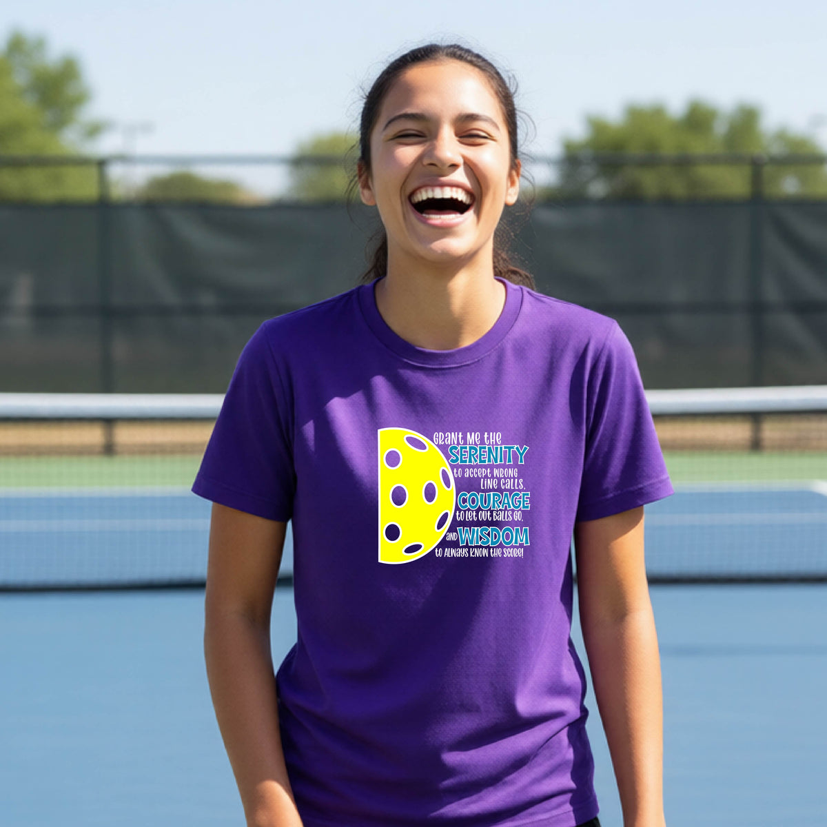 A young person on a pickleball court wearing a purple short-sleeve t-shirt with a graphic of the Serenity Prayer made for pickleball:  “Grant me the serenity to Accept Wrong Line Calls; Courage to Let Out Balls Go; and, Wisdom to Always Know the Score” next to a half-pickleball icon, standing on a pickleball court.